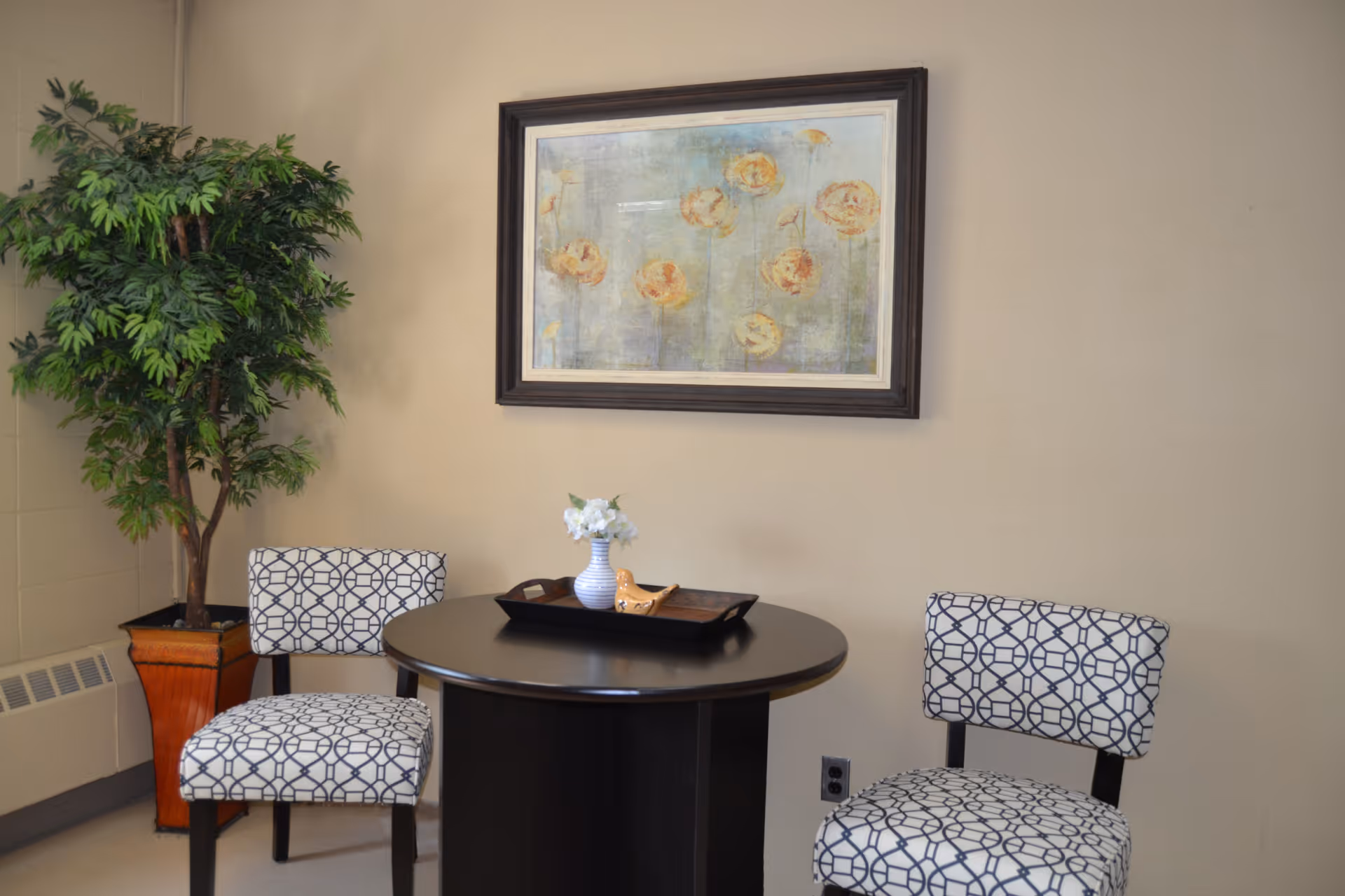A small seating area with a round black table and two patterned chairs with white and black geometric designs. On the table is a black tray holding a small white vase with white flowers and a decorative bird figurine. A large framed painting of yellow flowers hangs on the beige wall behind the table. To the left is a tall green potted plant in a brown square planter.