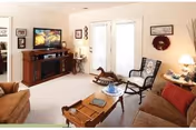 A cozy living room with beige walls and carpet, featuring a brown sofa with red cushions, a wooden coffee table with decorative items, a rocking chair, and a wooden entertainment center with a TV displaying a nature scene. French doors with glass panes lead outside, and the room is decorated with framed pictures and a wreath on the wall.
