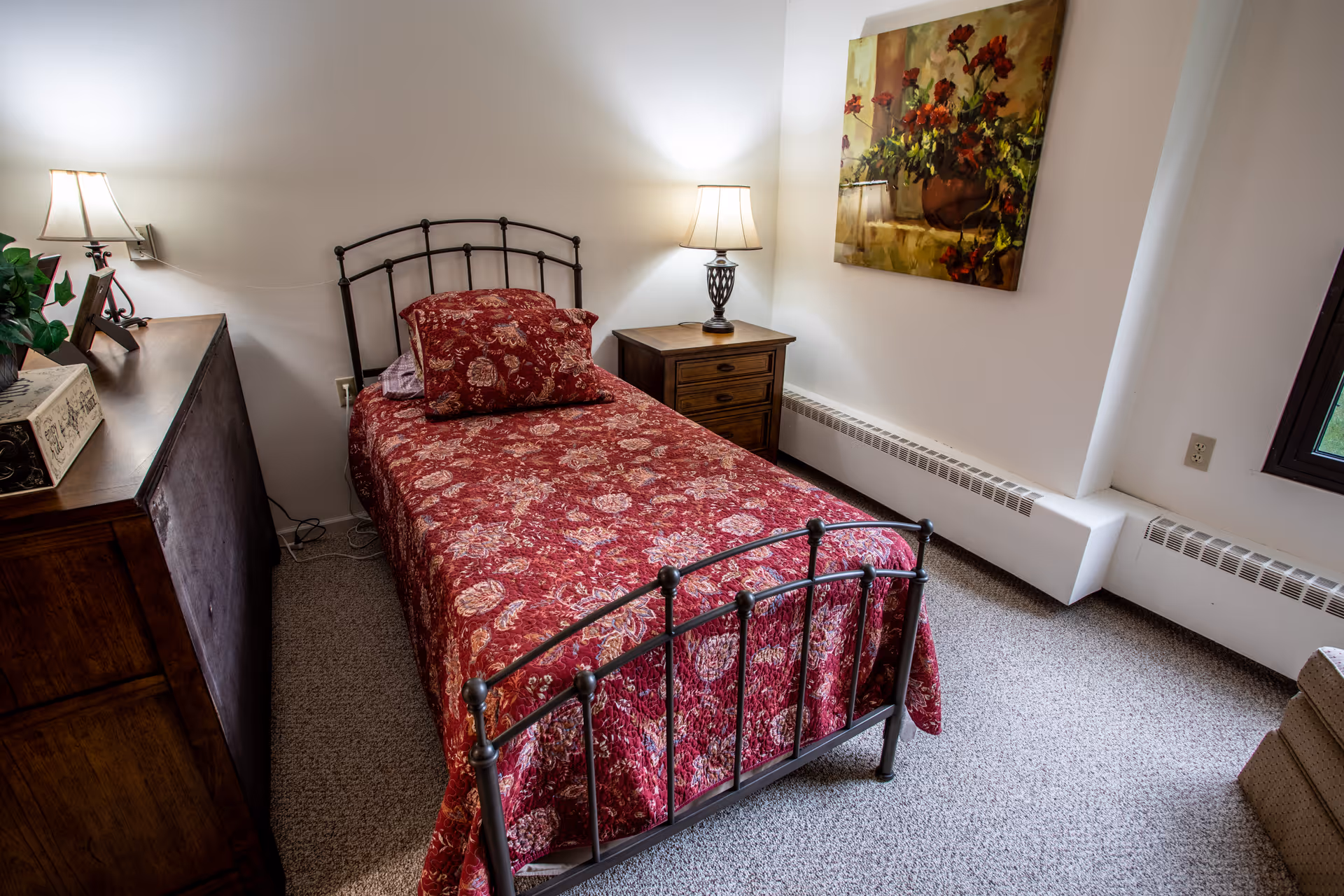 A cozy bedroom with a single metal bed frame covered in a red floral bedspread and matching pillow. There is a wooden nightstand with a lamp on it next to the bed, a wooden dresser with decorative items on the left, a painting of flowers on the wall, and a window letting in natural light. The room has carpeted flooring and white walls.