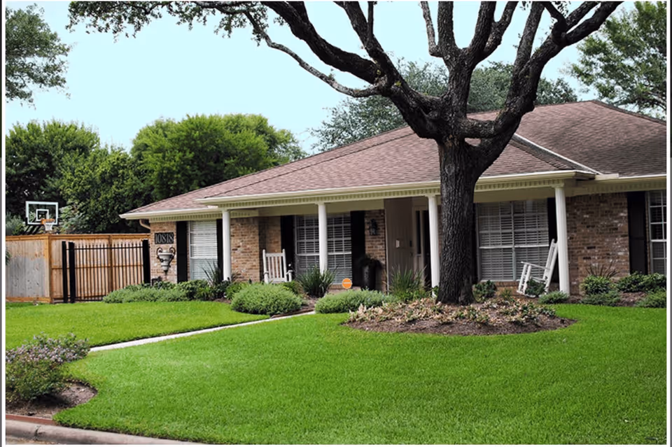 Front exterior view of a single-story brick building with a covered porch, white columns, rocking chairs, and a large tree in the front yard. The lawn is well-maintained with green grass and shrubs. A basketball hoop is visible near a wooden fence on the left side.