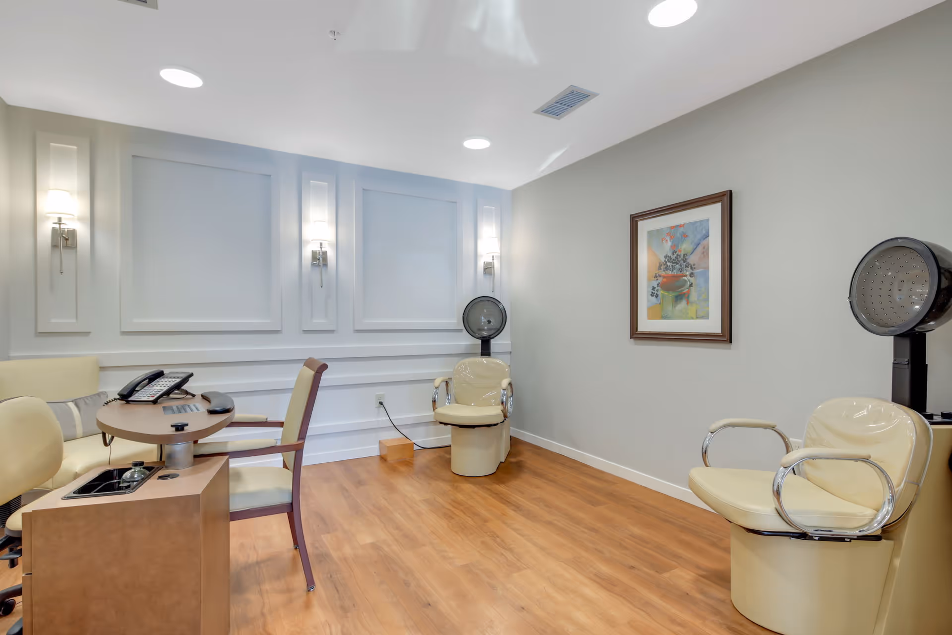 Interior of a salon room with two beige salon chairs equipped with hair dryers, a small round table with two chairs, a telephone, and a framed abstract painting on the wall. The room has wooden flooring, light gray walls with decorative paneling, and ceiling lights.