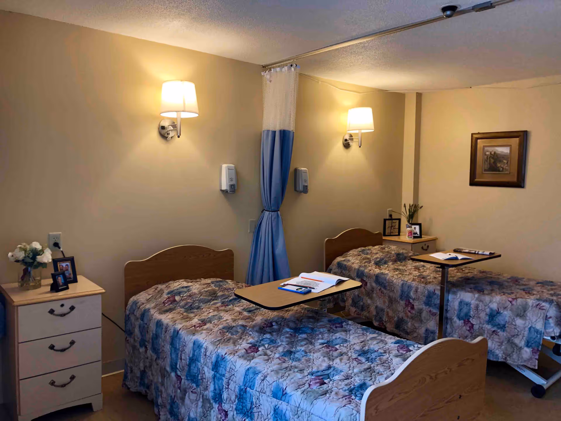 A room with two hospital-style beds, each with a floral patterned bedspread and an overbed table with books and papers. There are wall-mounted lamps above each bed, a blue privacy curtain between the beds, and nightstands with framed photos and flowers on either side. A framed picture hangs on the wall.