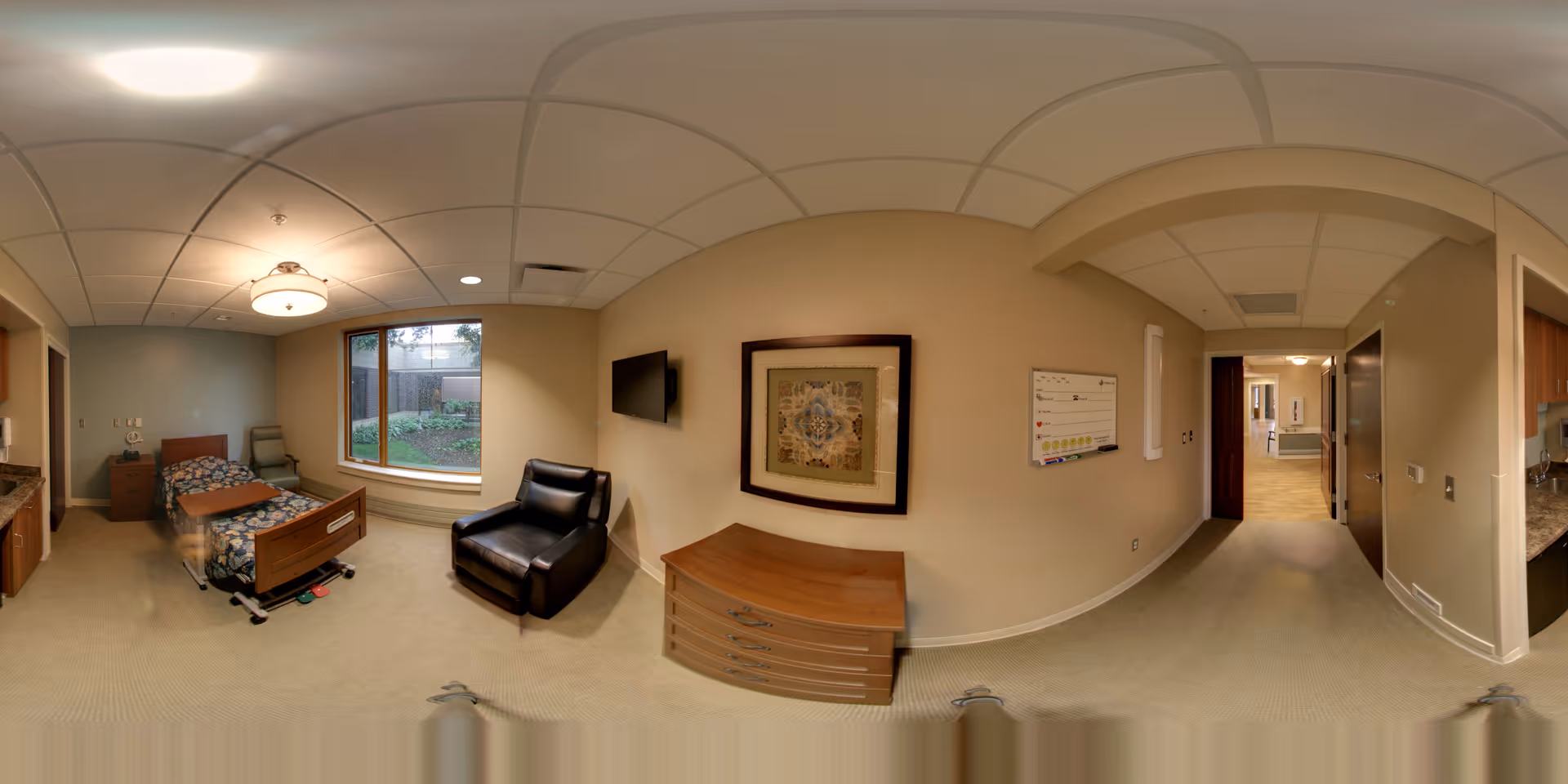 A senior living facility bedroom with a hospital-style bed, a bedside table, a comfortable black armchair, a large window overlooking a garden, a wall-mounted TV, a framed artwork, and a whiteboard on the wall. The room has beige walls and carpeted floor, with a hallway leading to other areas.