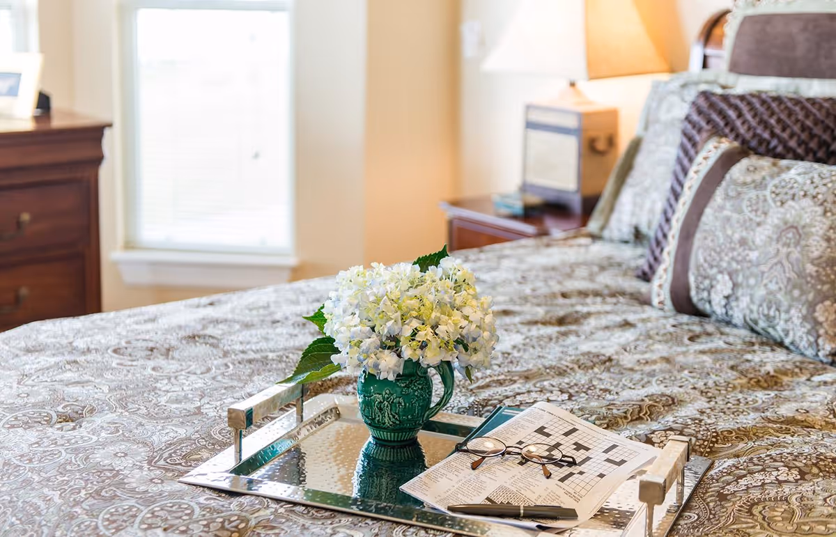 A neatly made bed with patterned bedding and multiple decorative pillows. On the bed is a silver tray holding a green vase with white flowers, a crossword puzzle, a pair of reading glasses, and a pen. In the background, there is a wooden nightstand with a lamp and a window with blinds.