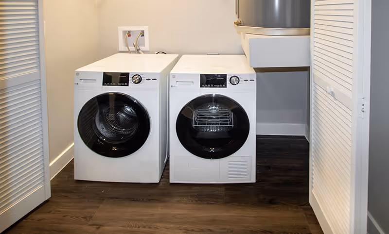 A laundry room with a white front-loading washing machine and dryer side by side on a dark wood floor, with white louvered doors on either side and a water heater mounted on the wall above the dryer.