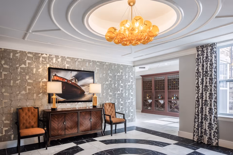 Bright, elegant lobby-style room with a central chandelier, patterned marble floor, a sideboard flanked by two chairs and lamps, artwork on a textured wall, and a wine cabinet in the background.