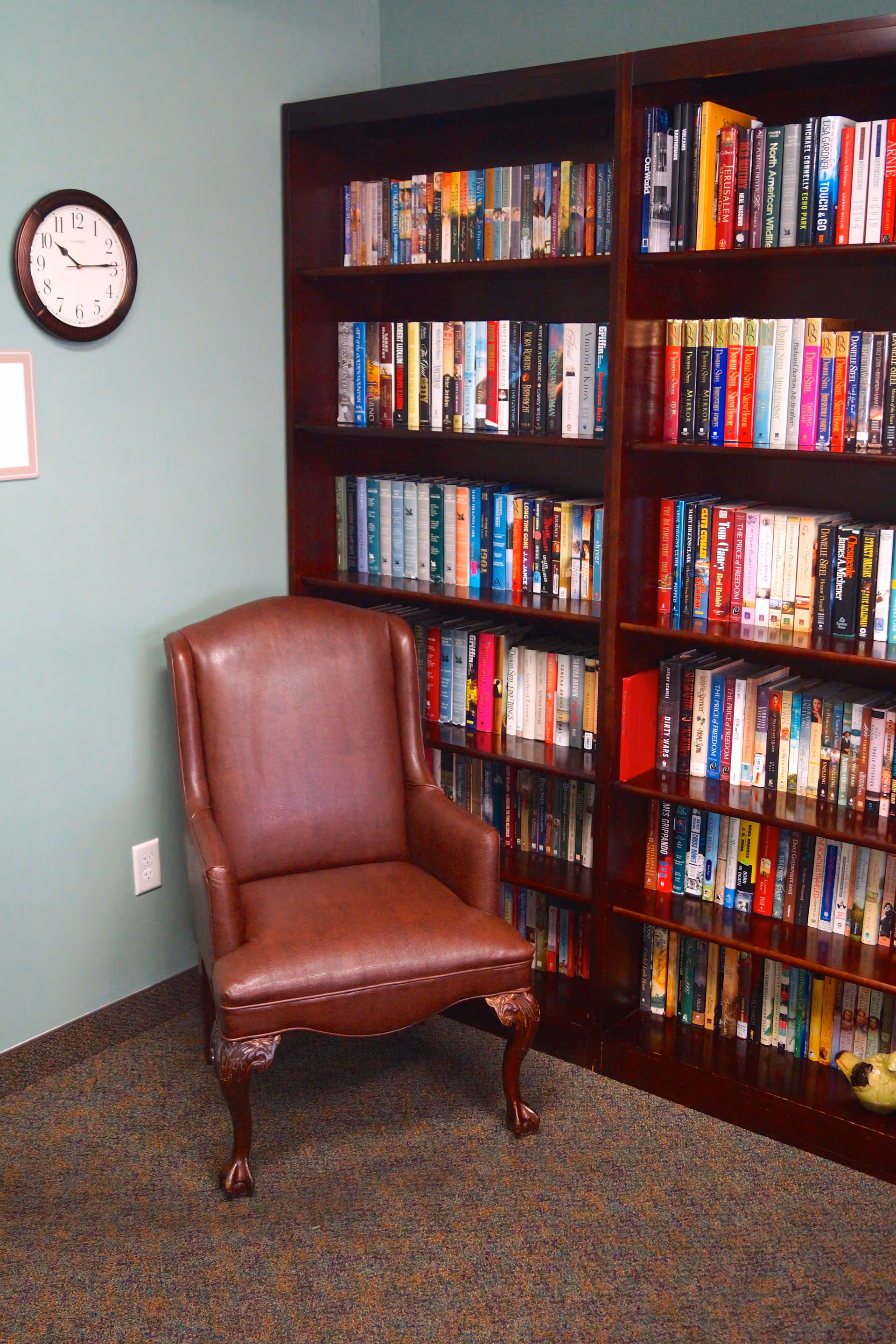 A cozy reading corner with a brown leather armchair placed in front of a tall wooden bookshelf filled with books. A round wall clock shows the time as 3:50. The walls are painted light green and the floor is carpeted.