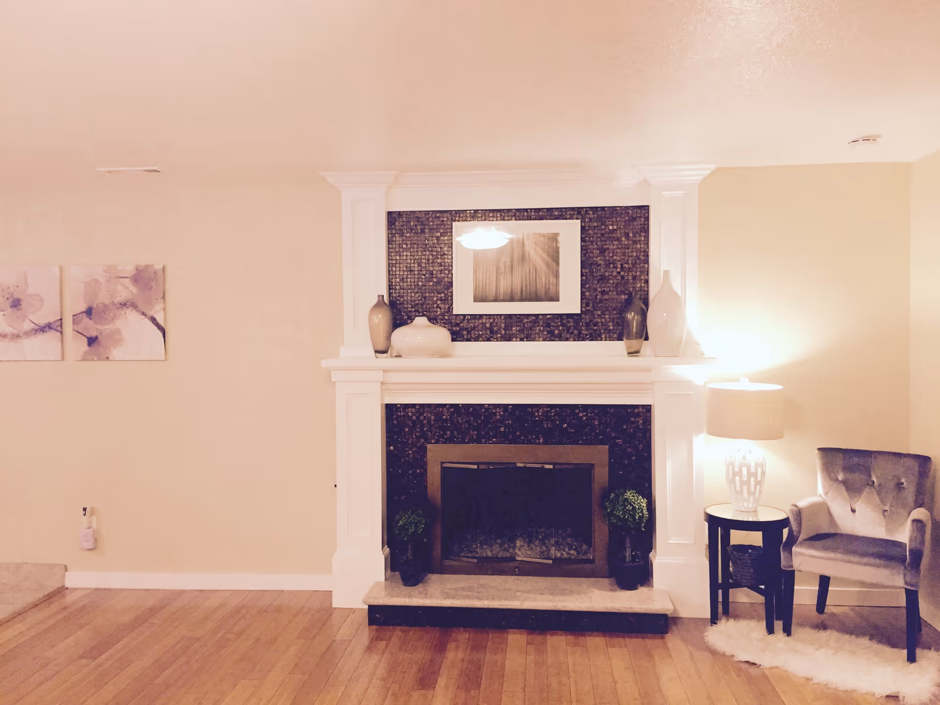 A cozy living room area featuring a white fireplace with dark mosaic tiles, decorated with vases and a framed picture above. To the right of the fireplace is a small round table with a lamp and a gray upholstered armchair on a white rug. The floor is wooden, and light-colored walls have two floral paintings on the left side.