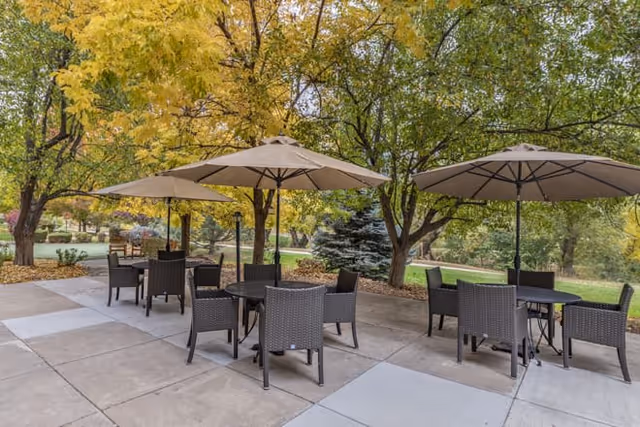 Outdoor patio area with several round tables and chairs under large beige umbrellas, surrounded by trees with green and yellow leaves.
