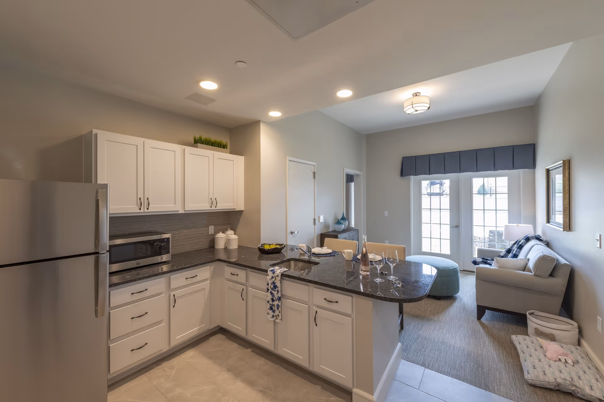 A modern kitchen and living room area in a senior living facility. The kitchen features white cabinets, a stainless steel refrigerator, a microwave, and a black granite countertop with a sink. The living room has a gray sofa, a blue ottoman, a side table with a lamp, and large glass doors with blue valances letting in natural light.