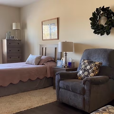 A cozy bedroom with a single bed covered in a mauve quilt and pillows, a wooden dresser with a lamp and decorative items, a nightstand with a lamp, framed photo, and tissue box, a comfortable gray armchair with a patterned cushion, and a green wreath hanging on the beige wall.