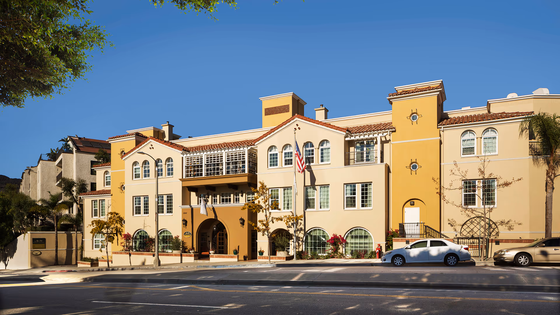 Exterior front view of a multi-story senior living facility building with beige and yellow walls, red tile roof, multiple windows, an American flag, and parked cars along the street in front.