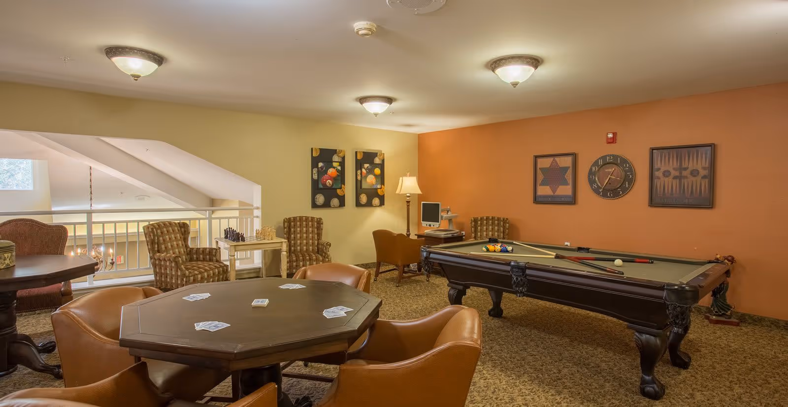 A recreational room with a pool table, a card table with playing cards, several armchairs, a computer desk with a lamp, and wall decorations including a clock and framed artwork.