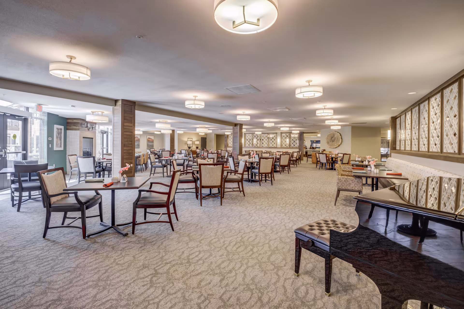 Spacious dining room in Merrill Gardens at Brentwood with multiple tables and chairs arranged neatly. The room features soft lighting from ceiling fixtures, a carpeted floor, decorative wall panels, and a grand piano in the foreground on the right side. Large windows and glass doors allow natural light to enter the space.