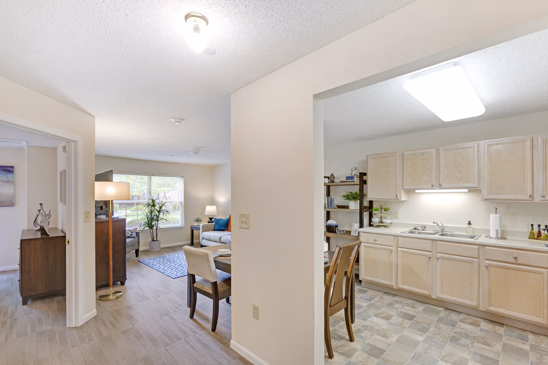 View of an interior living space in a senior living facility showing a kitchen with light wood cabinets and a dining area with a table and chairs. Adjacent to the kitchen is a living room with a large window, a sofa, a side table with a lamp, a floor lamp, and a potted plant. A hallway leads to another room with a wooden dresser and decorative items.