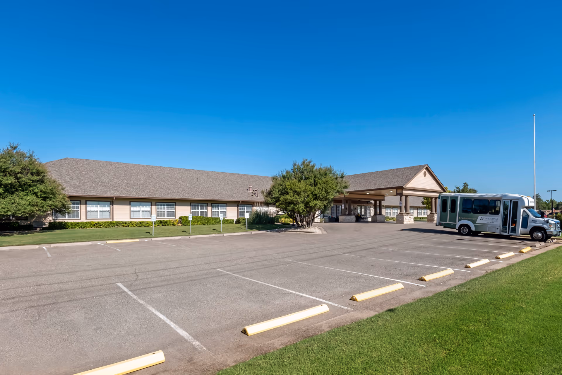 Exterior view of Brookdale Monterey senior living facility showing a single-story building with a covered entrance, a parking lot with marked spaces, a shuttle bus parked near the entrance, green grass, and clear blue sky.