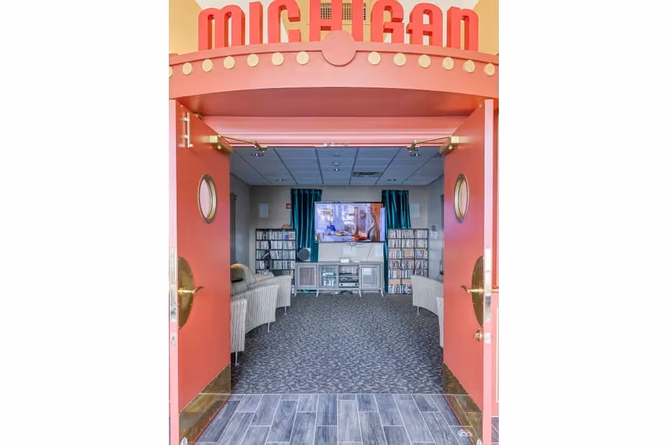 View through open red double doors into a cozy media room with patterned carpet, two striped armchairs facing a wall-mounted flat screen TV, and shelves filled with DVDs or CDs on either side of a media console. The top of the doorway has a decorative sign that reads 'MICHIGAN'.