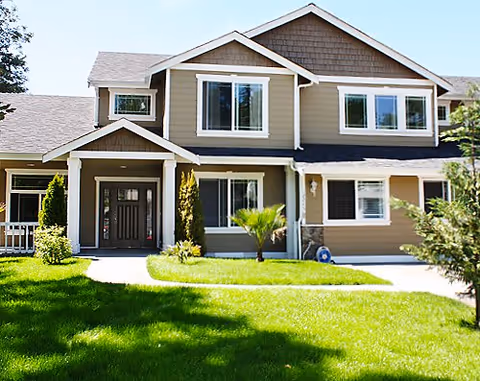 Front exterior view of a two-story residential-style building with beige siding, white trim, multiple windows, a dark front door, and a well-maintained green lawn with small trees and shrubs.