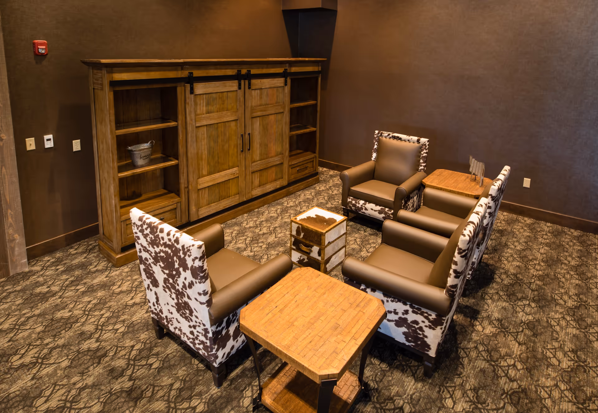 A cozy seating area with four armchairs featuring brown and white cowhide upholstery arranged around two small wooden tables in a room with brown walls and patterned carpet. A large wooden cabinet with sliding doors and open shelves is against the wall.