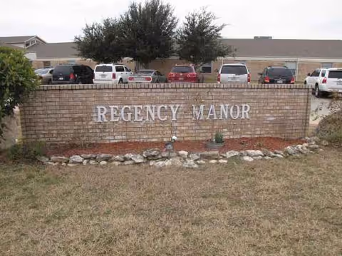Brick sign reading Regency Manor with parked cars behind and a building.
