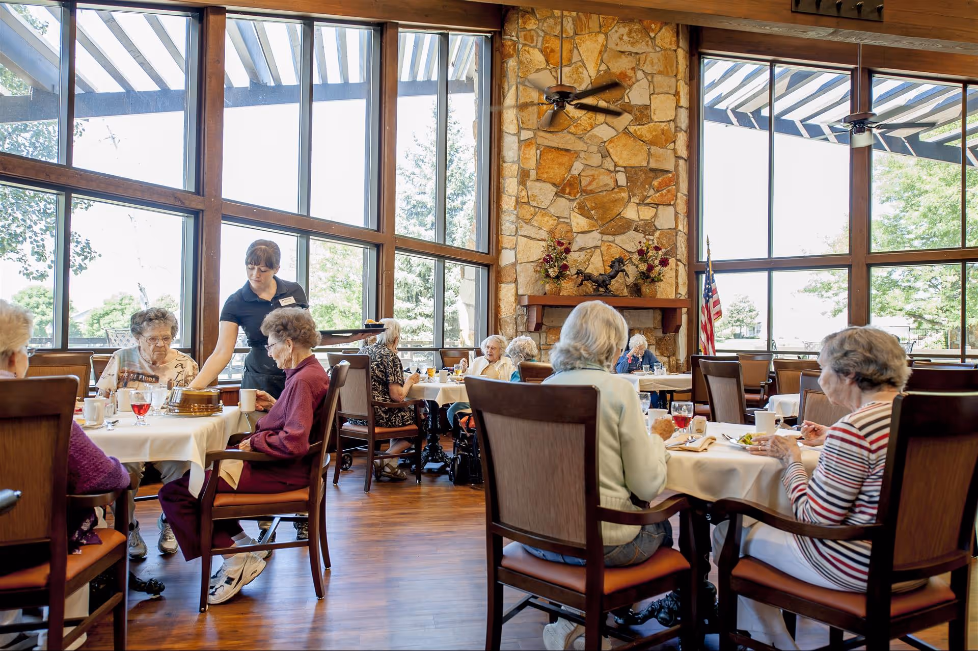A dining room in an assisted living facility with elderly residents seated at tables covered with white tablecloths, eating and drinking. A staff member is serving food to one of the residents. The room has large windows with wooden frames, a stone fireplace, ceiling fans, and an American flag near the window.