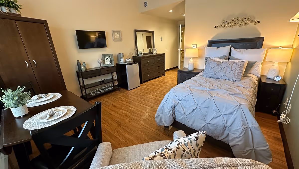 A cozy bedroom in a senior living facility featuring a neatly made bed with gray bedding, two bedside tables with lamps, a dresser with a mirror, a small table with two place settings, a mini refrigerator, a wall-mounted TV, and a comfortable armchair with a floral pillow. The room has wooden flooring and warm lighting.
