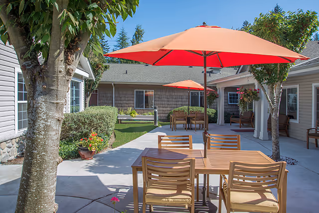 Sunlit courtyard patio with wooden tables and chairs under red umbrellas between single-story residential buildings.