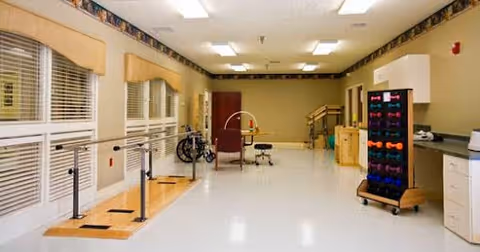Spacious indoor therapy room with parallel walking bars, exercise equipment, a rack of hand weights, chairs and a wheelchair.