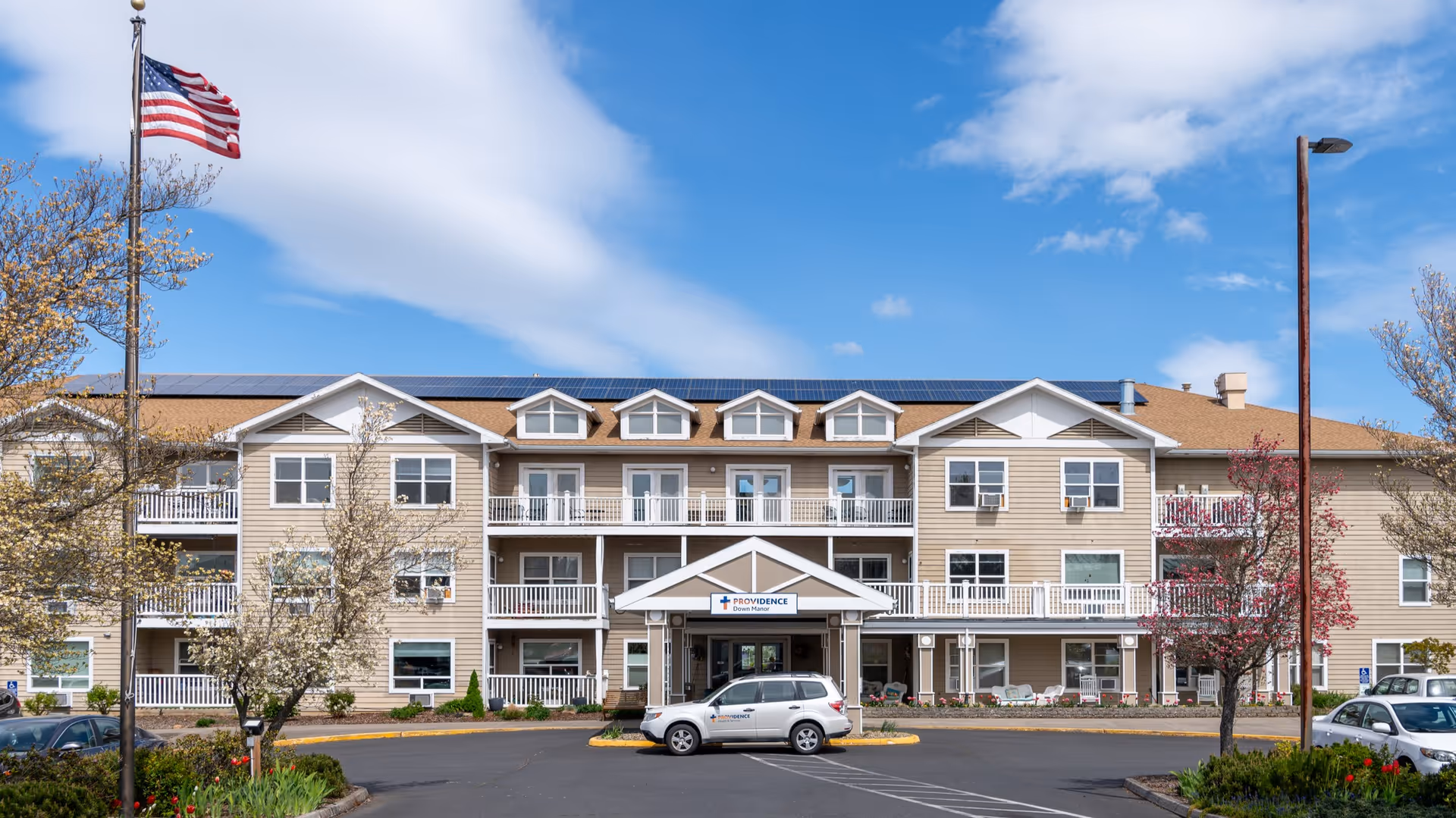 Front exterior view of Providence Down Manor - Hood River, a three-story senior living facility with beige siding, white trim, balconies, and a peaked entrance canopy. An American flag flies on a flagpole to the left, and several cars are parked in front of the building under a partly cloudy blue sky.