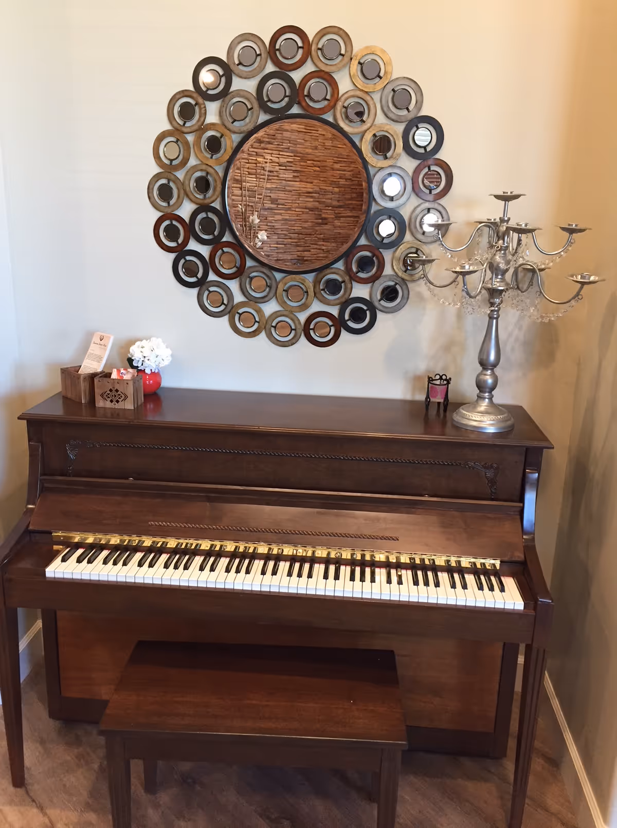 Upright wooden piano with a bench, topped with decorative items and a large circular multi-ring mirror on the wall above.