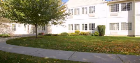 Two-story white senior living building with green lawn, tree, and curved sidewalk