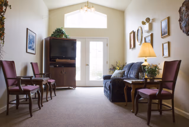 A cozy living room area with a blue patterned sofa, a wooden TV cabinet with a television, and several wooden chairs with red cushions. The room has beige walls adorned with framed pictures and a wall lamp. A table lamp on a side table next to the sofa provides warm lighting. There is a large window and glass double doors letting in natural light at the far end of the room.