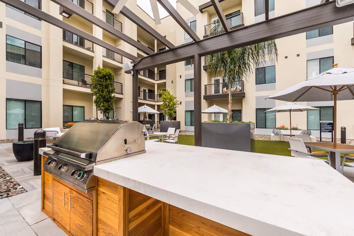 Outdoor courtyard with a built-in stainless steel grill and countertop, patio seating with umbrellas, pergola, and surrounding apartment balconies.