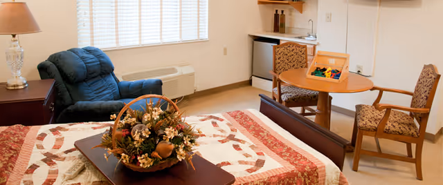 A cozy senior living bedroom featuring a bed with a patterned quilt and a floral centerpiece on a wooden tray. The room includes a blue recliner chair, a wooden side table with a lamp, a small round table with two upholstered chairs, a mini refrigerator, and a small sink under a wall-mounted cabinet. A window with blinds allows natural light into the room.
