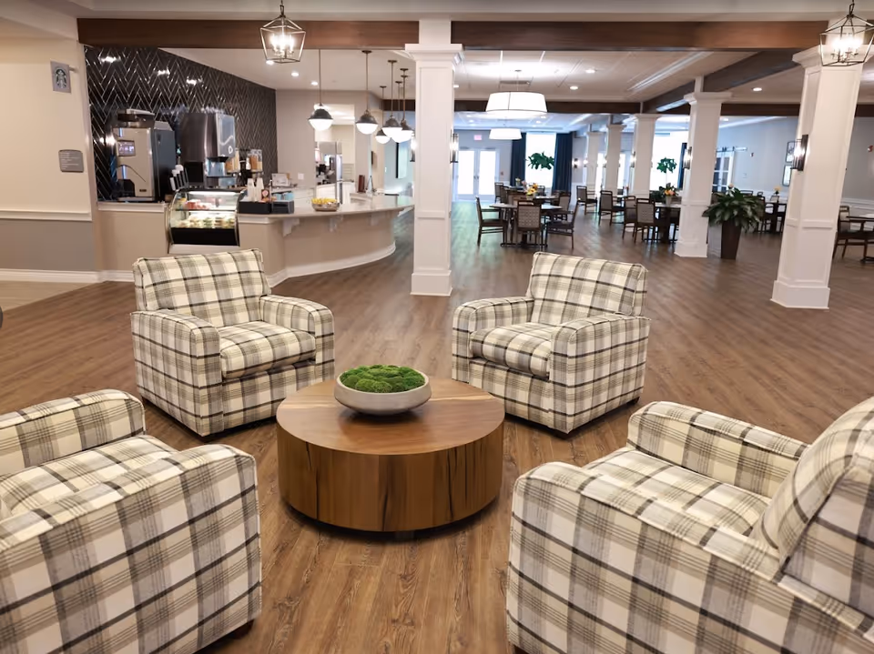 A spacious and well-lit common area in a senior living facility featuring four plaid upholstered armchairs arranged around a round wooden coffee table with a decorative bowl of greenery. In the background, there is a dining area with tables and chairs, pendant lights, and a counter with a coffee machine and display case.