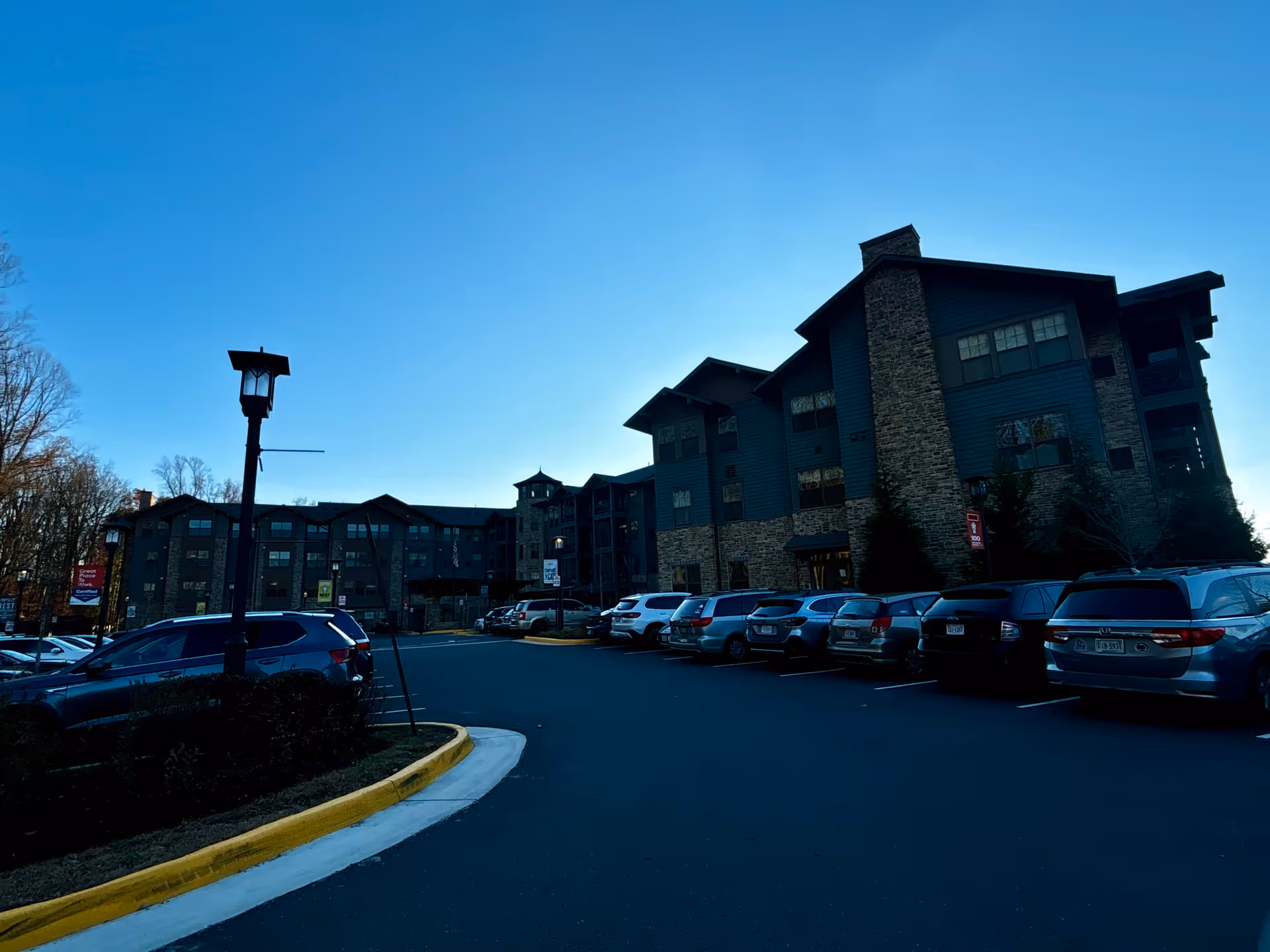 Exterior view of a large senior living facility building with multiple floors, stone and wood facade, and a parking lot filled with cars in front. The sky is clear and blue.