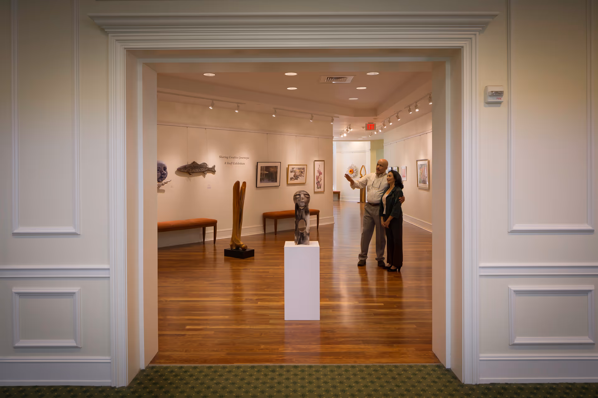 An art gallery room with wooden floors and white walls displaying various sculptures and framed artworks. Two people stand near the center, looking at the art and pointing. The room is viewed through a wide doorway with white trim and molding.