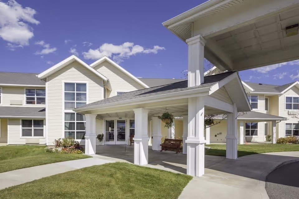 Exterior view of The Springs at Butte senior living facility showing a covered entrance with white pillars, a wooden bench swing, and a well-maintained lawn under a blue sky with scattered clouds.