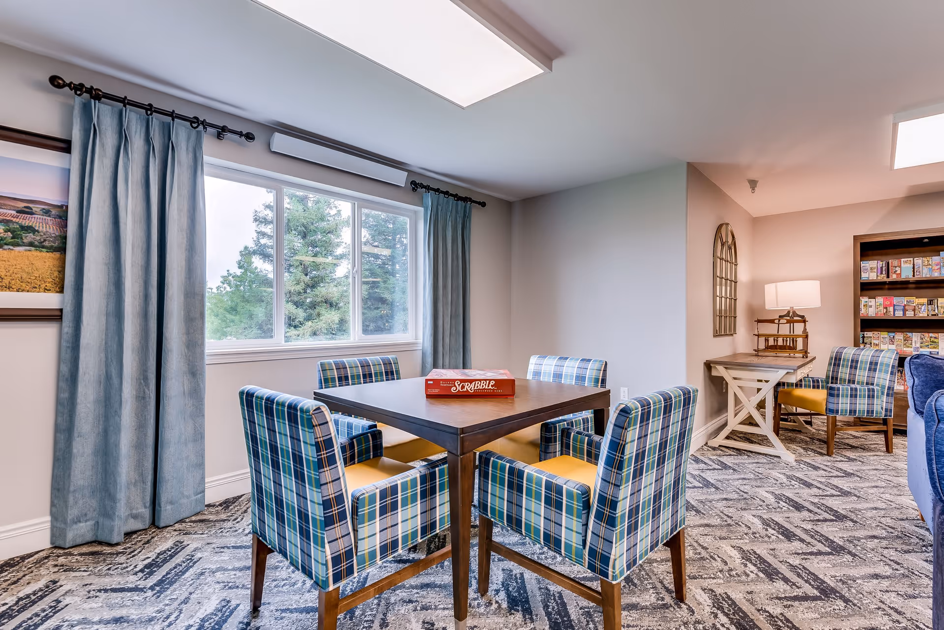 A cozy common area in a senior living facility featuring a square wooden table with four plaid upholstered chairs around it. A Scrabble board game is placed on the table. The room has a large window with blue curtains, a patterned carpet, a side table with a lamp, and a bookshelf filled with board games and books.