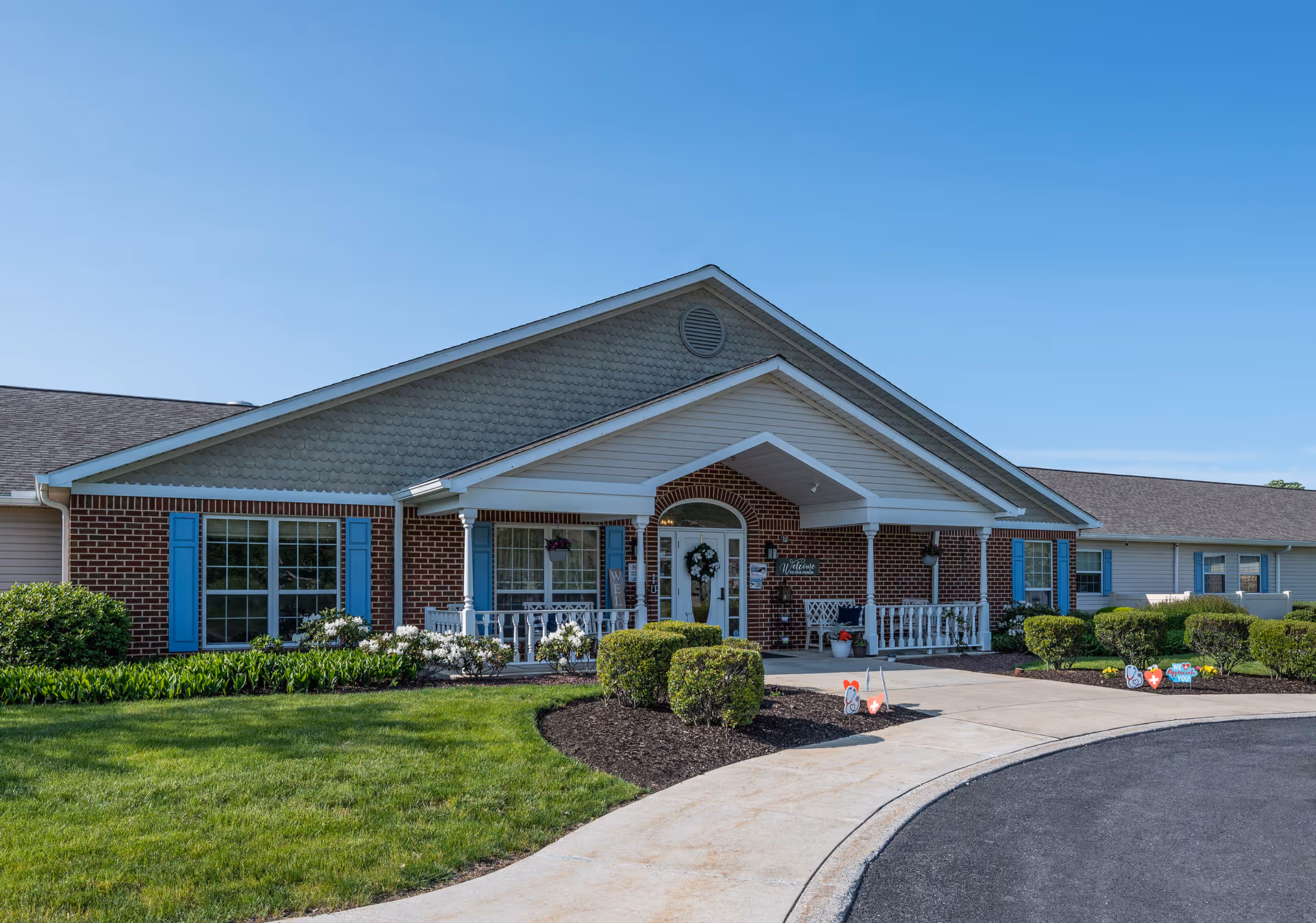 Single-story brick and siding memory care facility with a covered front porch, landscaped lawn and circular driveway under a clear blue sky.