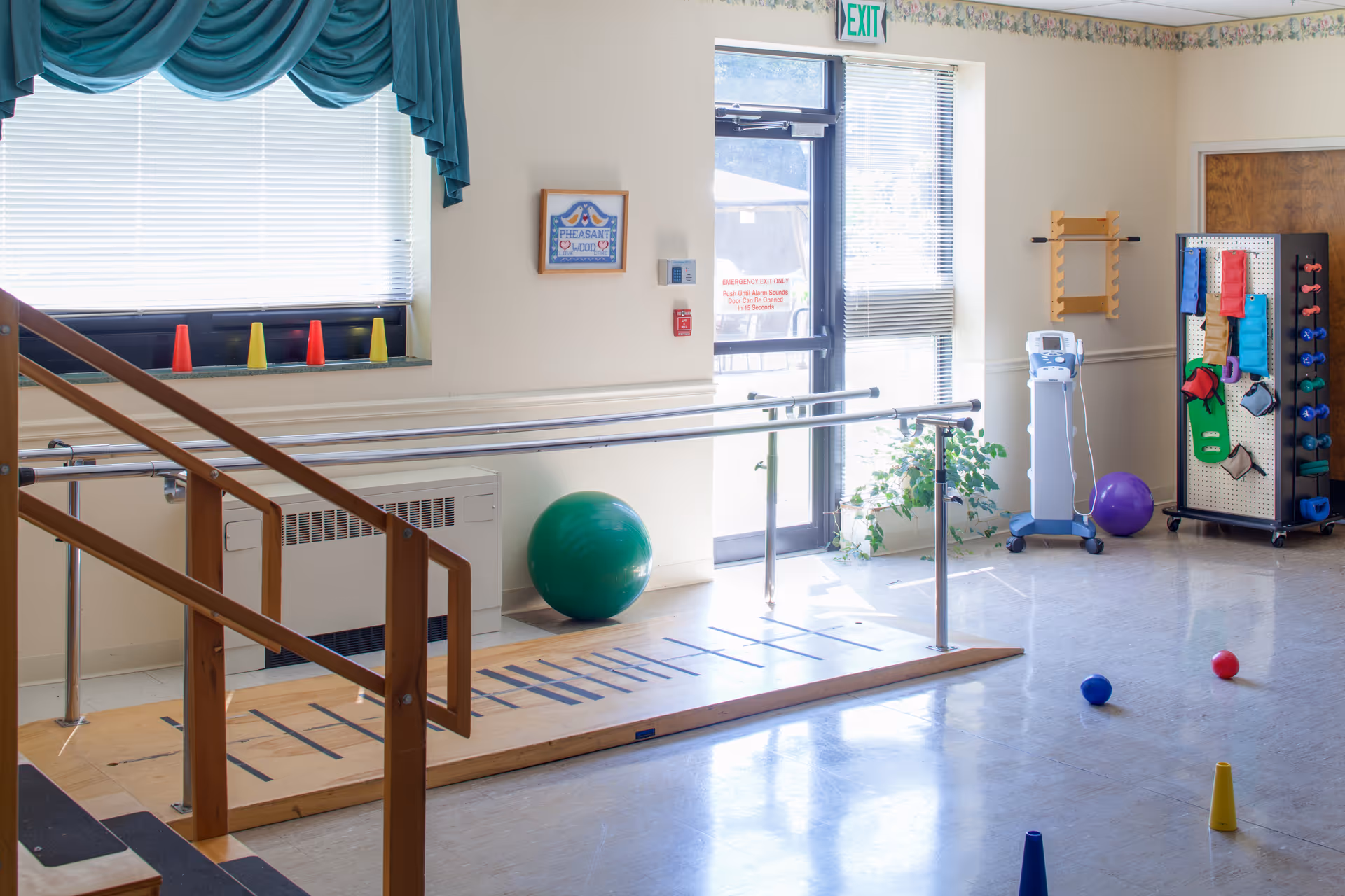A bright physical therapy room with parallel bars for walking exercises, colorful cones and balls scattered on the floor, a green exercise ball, and various therapy equipment on racks. A window with teal curtains and a door with an exit sign are visible.
