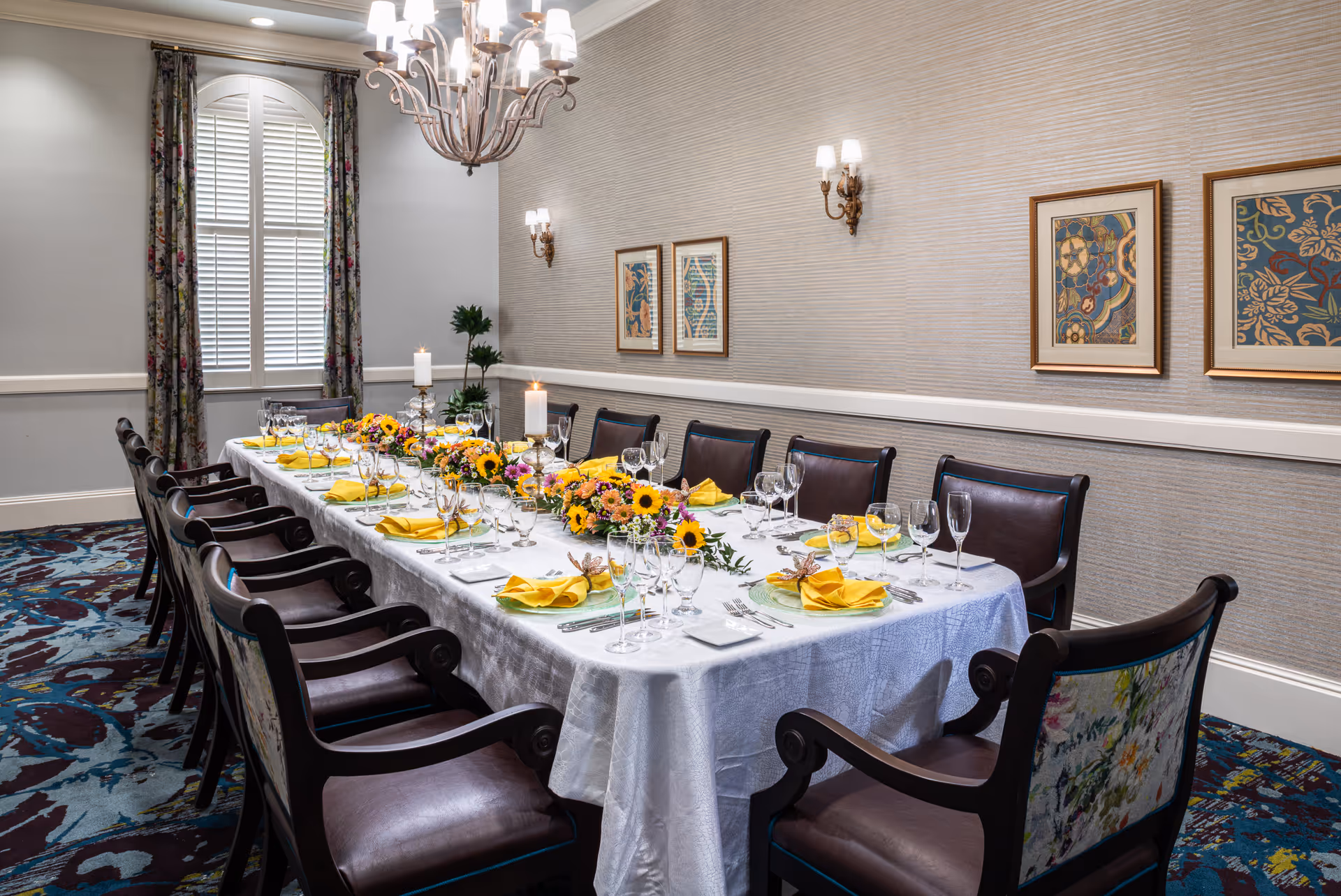 A formal dining room with a long rectangular table set for a meal. The table is covered with a white tablecloth and decorated with yellow napkins, glassware, silverware, and floral centerpieces featuring sunflowers. The room has patterned carpet, floral curtains framing a window with white shutters, wall sconces, framed artwork, and a chandelier hanging above the table.
