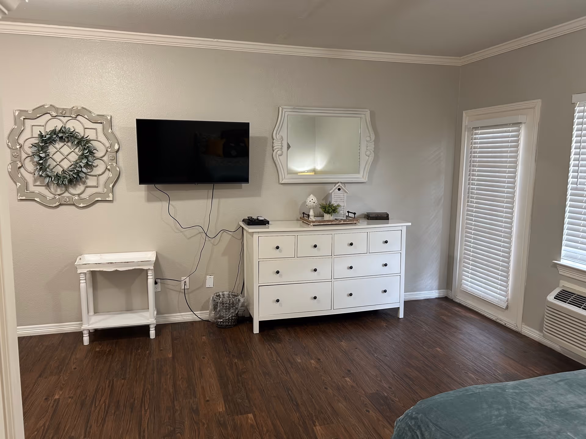 A room with wooden flooring featuring a white dresser with multiple drawers, a decorative mirror above it, and a mounted flat-screen TV on the wall. To the left of the dresser is a small white side table and a decorative wall piece with a green wreath. Two windows with white blinds are on the right side of the room, and a portion of a bed with a green blanket is visible in the lower right corner.