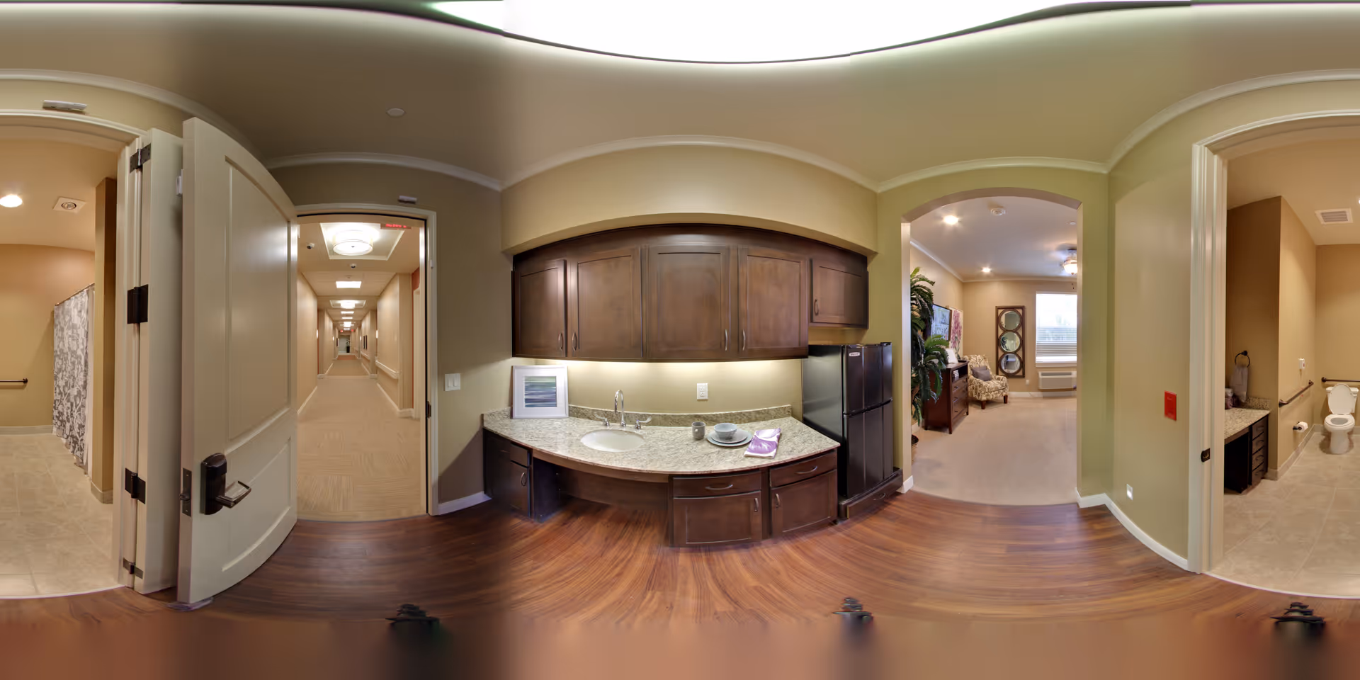 A panoramic view of an interior space in a senior living facility showing a kitchenette with dark wood cabinets, a sink, countertop, and a small refrigerator. To the left, there is an open door leading to a hallway and a bathroom with a shower curtain visible. To the right, there is another open door leading to a bathroom with a toilet and sink. Beyond the kitchenette, an arched doorway opens into a living room area with a chair, plants, and windows letting in natural light.