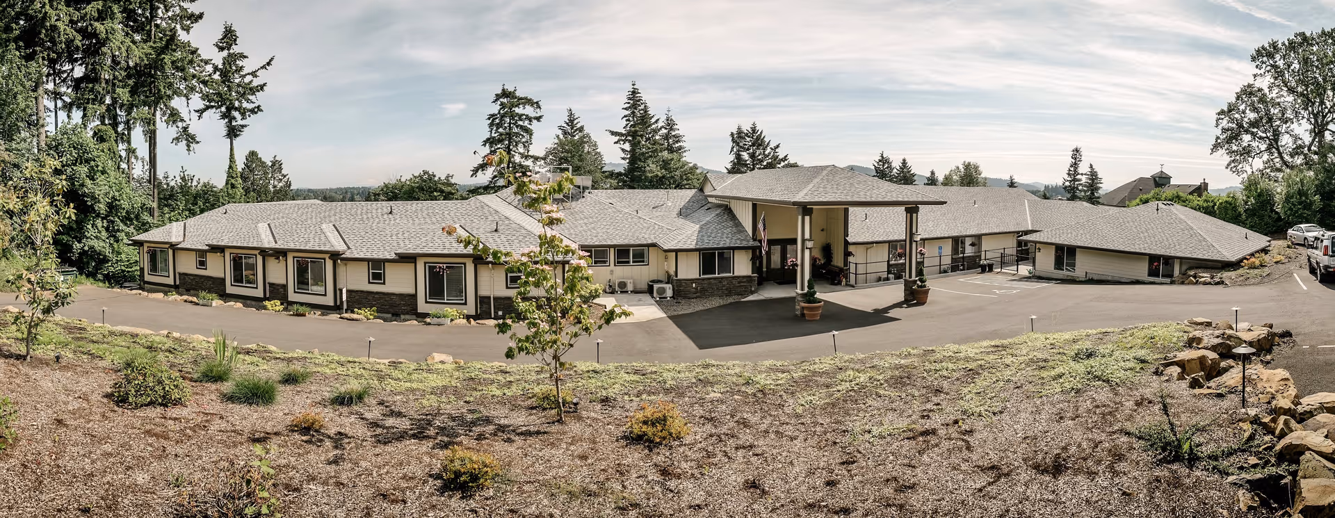Wide exterior view of Clackamas Heights Senior Living facility showing a single-story building with multiple windows, a covered entrance with pillars, surrounded by trees and landscaping under a partly cloudy sky.