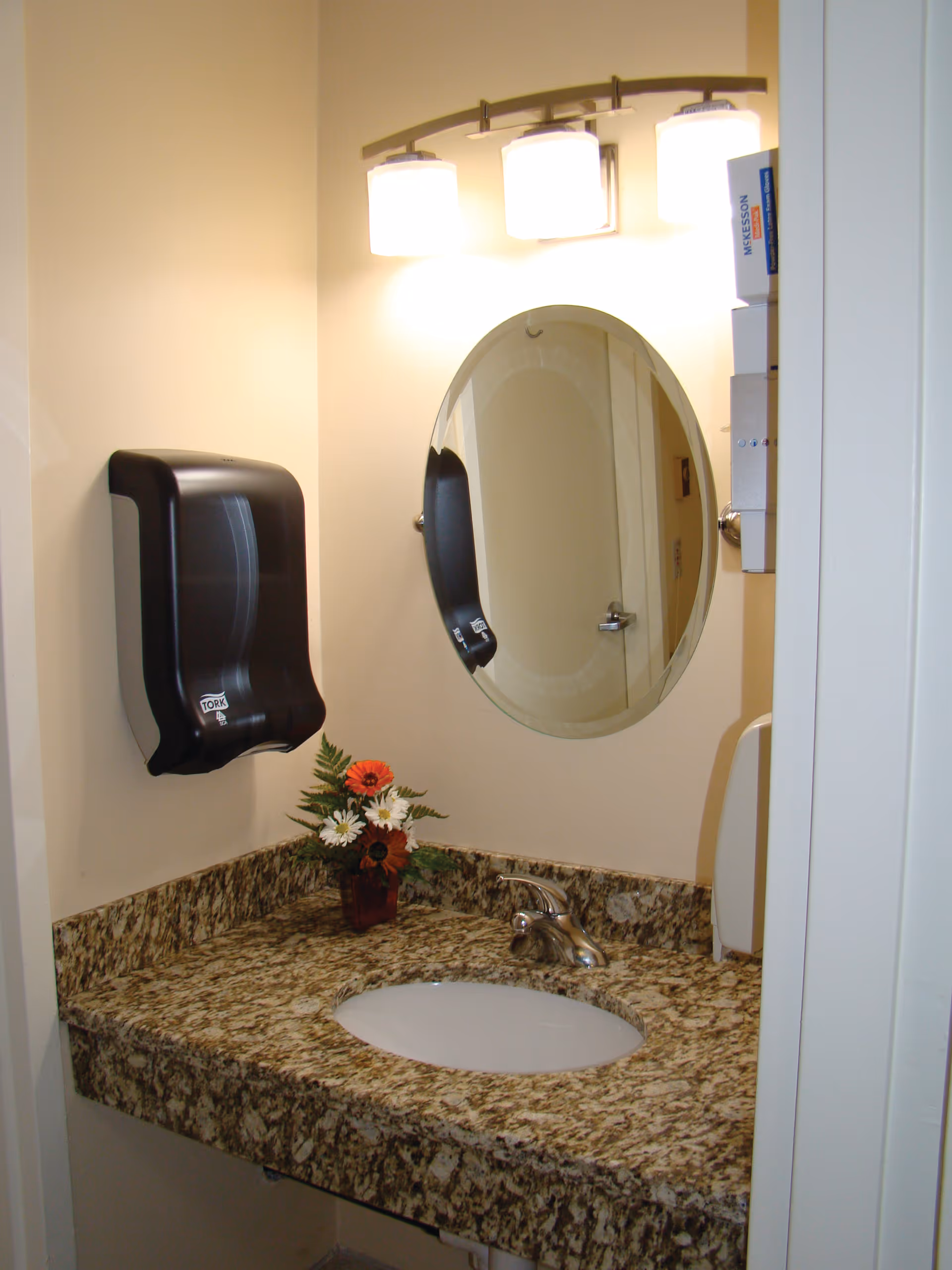 A bathroom sink area with a granite countertop, an oval mirror above the sink, a three-light fixture, a black paper towel dispenser mounted on the wall, a small vase with flowers on the counter, and a soap dispenser on the right side.