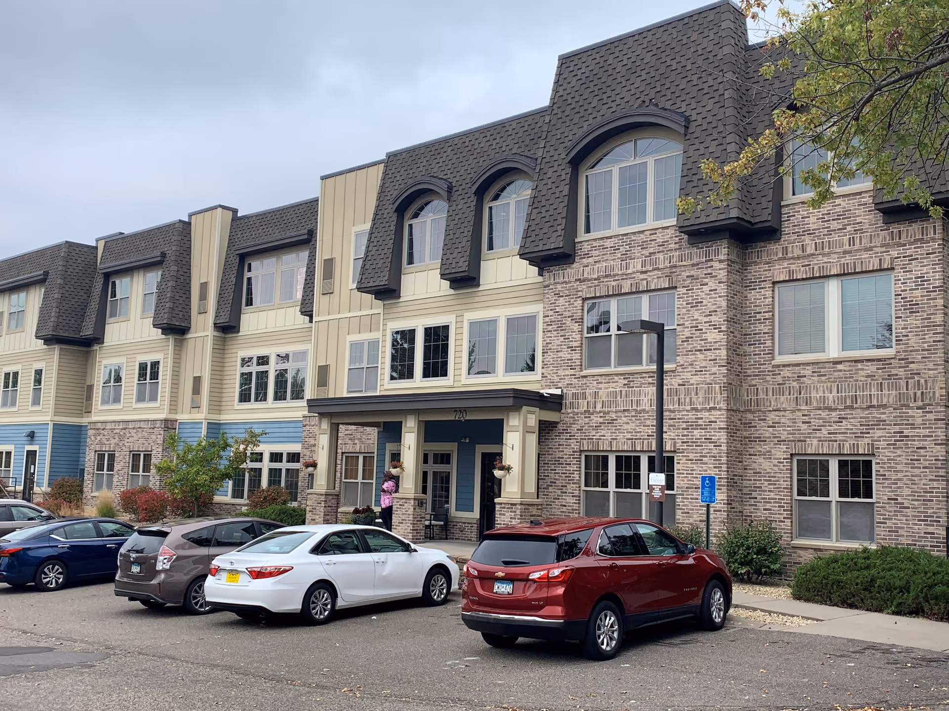 Exterior view of a three-story assisted living and memory care facility with a combination of brick and siding facade. Several cars are parked in front of the building near the entrance, where a person is standing. The sky is overcast and some trees with autumn leaves are visible.
