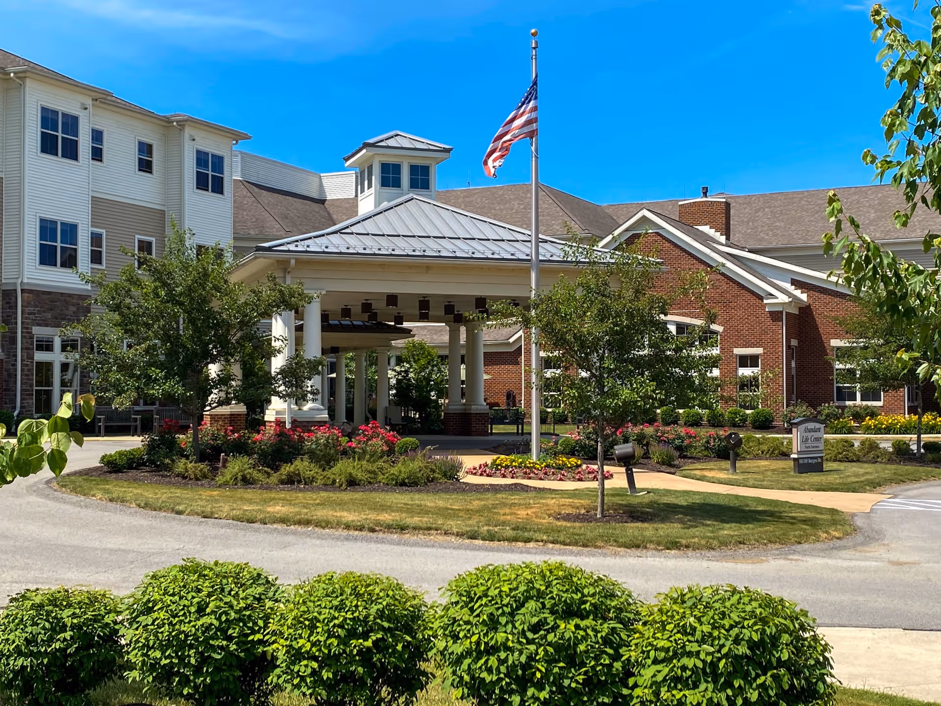 Exterior view of Newhaven Court Personal Care Apartments of Passavant Community showing a large building with white and brick walls, a covered entrance with white columns, an American flag on a flagpole, well-maintained landscaping with bushes, trees, and flowers, and a clear blue sky.