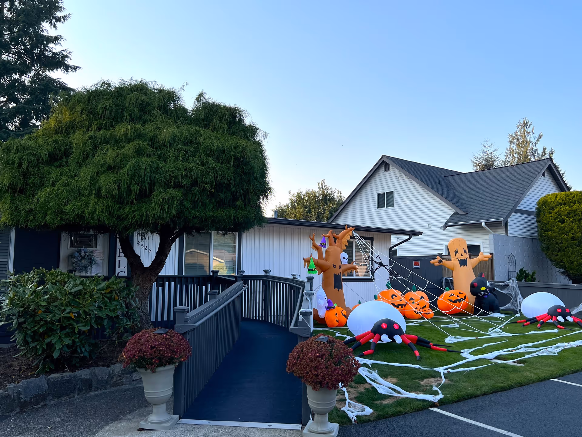 Exterior view of a senior care home decorated with Halloween inflatables including pumpkins, spiders, a black cat, and spooky trees on a green lawn next to a ramp leading to the entrance. There are potted flowers on either side of the ramp and a large tree on the left side of the building.