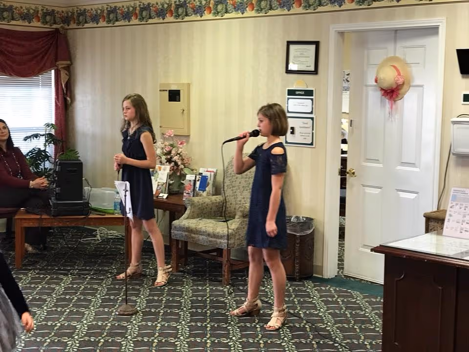 Two young girls in navy blue dresses are performing with microphones in a carpeted room with floral wallpaper border. A woman is seated on a wooden bench to the left, and there is a patterned armchair, a table with flowers and brochures, and a white door with a decorative hat hanging on it in the background.